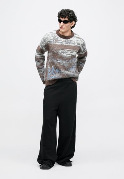 Pleteni pulover z brazimi, bež in modrimi odtenki, z okrolim izrezom in rebrastimi manšetami, kombiniran z ohlapnimi črnimi hlačami in obutvijo.