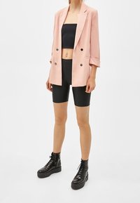 Ljusrosa dubbelknäppt kavaj över en svart cropped topp och svarta cykelshorts, matchat med kraftiga svarta snörkängor.