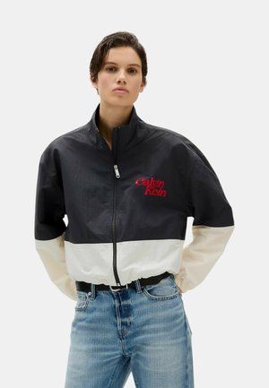 Giacca a windbreaker bicolore in bianco e nero con colletto alto e logo rosso. Presenta una chiusura con zip anteriore e polsini elasticizzati.