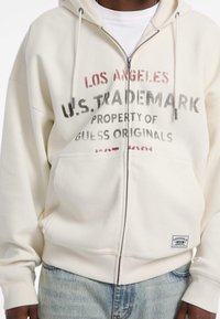 Cremefarbene Zip-Hoodie aus strukturiertem Stoff, mit rot-schwarzem grafischen Text. Rippbündchen und -bund, mit einem Logopatch am Saum.