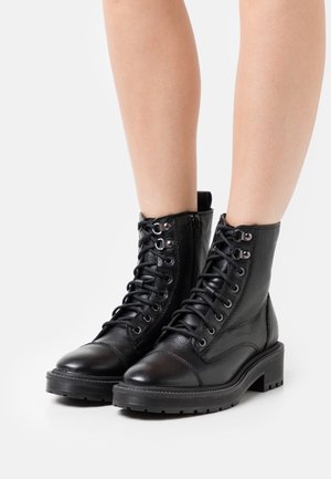 Schwarze Leder-Schnürstiefeletten, getragen an nackten Beinen vor weißem Hintergrund.