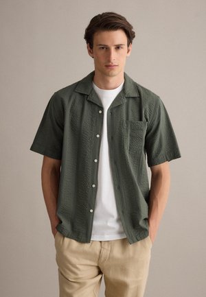 Jonge man draagt een gestructureerd olijfgroen overhemd met korte mouwen over een wit T-shirt en beige broek, staand tegen een effen achtergrond.