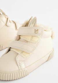 Beige children's shoes with velcro straps, textured fur trim, and bunny ear accents. Made from synthetic material with a rubber sole.