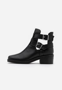 Bottines noires en cuir avec un bout pointu, des sangles à double boucle et un talon carré. Texture lisse et détails découpés sur les côtés.
