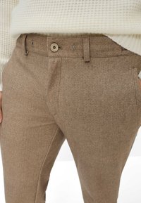 Bruine broeken gemaakt van een textuurstof. Voorzien van een knoopsluiting, zijzakken en een getailleerde pasvorm. Het ontwerp lijkt strak en modern.
