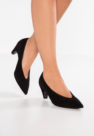 Femme portant des chaussures en daim noir à talon moyen et bout pointu, jambes croisées aux chevilles sur un sol et un fond blancs.