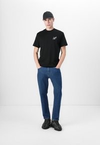 Svart bomull t-skjorte med logo, kombinert med blå denimjeans og sorte joggesko. Modellen har på seg en svart caps og står med hendene i lommene.