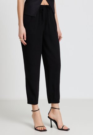 Femme portant un jogging noir élastique au niveau des chevilles, un haut noir sans manches, des bagues dorées et des sandales noires à talons hauts avec des brides, debout sur un fond uni.