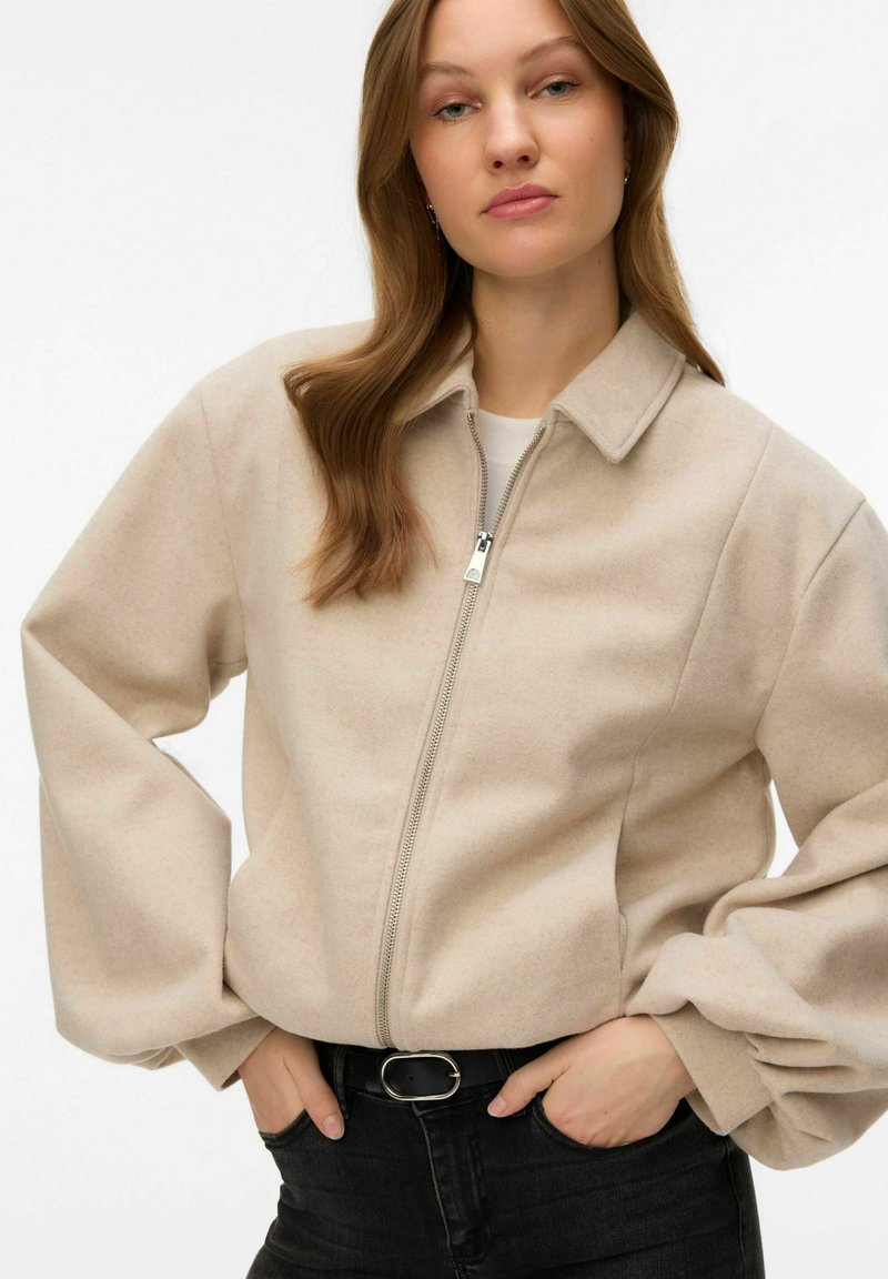 Veste beige à fermeture éclair avec un col en forme de chemise et des manches volumineuses, en tissu doux. Portée sur un haut blanc, associée à un jean foncé.