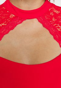 Haut rouge avec des détails en dentelle autour de l'encolure et une découpe en forme de trou de serrure laissant apparaître la peau du haut de la poitrine.