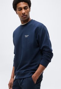 Άνδρας που φοράει μπλε σκούρο φούτερ και παντελόνι, με τη λέξη "marcopolo denim" στο στήθος του φούτερ, κοιτάζει προς τα κάτω με το ένα χέρι στην τσέπη.