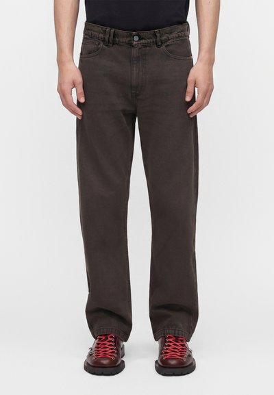 Homme portant un pantalon droit marron foncé et des chaussures de randonnée rouge foncé avec des lacets rouges vifs, debout devant un fond uni.