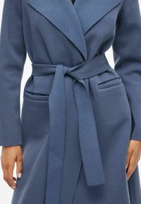 Cappotto blu avvolgente realizzato in tessuto morbido, caratterizzato da un ampio colletto, una cintura annodata e due tasche frontali, con linee pulite e una texture liscia.