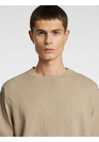 Beige strikkegenser med rund hals, ribbekant og myk tekstur, båret av en person med kort hår og øredobber.