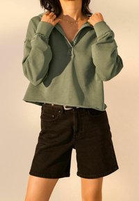 Groene trui, zachte stof, kraag met een patte, cropped fit, gecombineerd met zwarte spijkershorts, ruwe zoomdetails en zilverkleurige hardware.