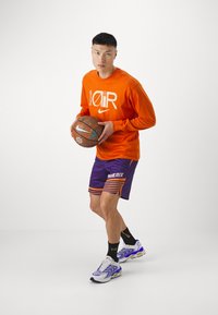 Pomarańczowa koszulka Nike z długim rękawem i białym logo, fioletowe szorty z pomarańczowymi paskami, czarne skarpetki i kolorowe buty sportowe, trzymający piłkę koszykową.