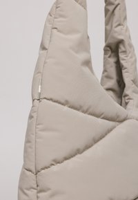 Borsa puffer beige chiaro con design trapuntato, caratterizzata da cuciture diagonali e una superficie liscia. Due manici robusti e imbottiti.