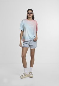 Nadměrné tričko s přechodem barev od modré k růžové, doplněné světle-denimovými šortkami. Nohy v ozdobných sandálech s bílým detailem ponožek.