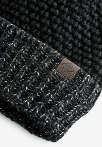 Mustra ja musta lõngaga kudumine beanie. Äär on ribadega musta ja halli segus, millel on pruun nahast logopatch.