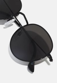 Schwarze Sonnenbrille mit übergroßen runden Gläsern, schwarzem Metallrahmen, klaren Nasenpads und schlanken Bügeln mit dezentem Branding-Detail.