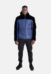 Giacca imbottita in blu navy e nero, con colletto alto, tasche con zip e una texture liscia. Abbinata a pantaloni cargo neri e sneakers.