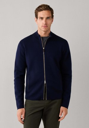 Homme portant un pull zippé bleu marine foncé par-dessus une chemise grise et un pantalon vert olive foncé, debout devant un fond gris clair uni.