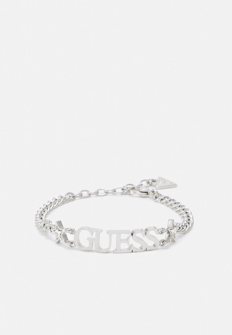 Bracelet chaîne en argent avec "GUESS" lettres et petits charms en forme d’étoile de chaque côté, posé à plat sur un fond blanc.