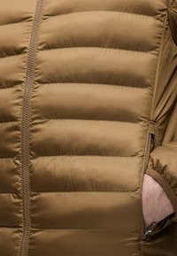 Brusthohe Nahaufnahme einer braunen gesteppter Jacke mit horizontaler Polsterung und einer Hand teilweise in der seitlichen Reißverschlusstasche.