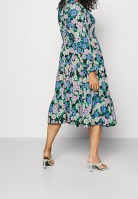 Robe midi florale avec un fond noir, ornée de fleurs violettes, bleues et vertes. Manches longues, design à volants et silhouette fluide.