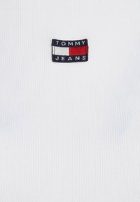 Bílá žebrovaná košile s hranatou vyšívanou nášivkou se textem "TOMMY JEANS" a designem v červené, bílé a modré barvě.