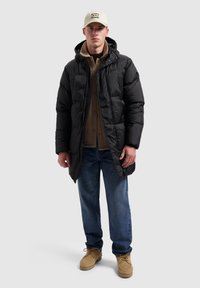 Zwarte pufferjas met capuchon, doorgestikt ontwerp, gecombineerd met een beige trui. Blauwe spijkerbroek en beige laarzen maken de outfit compleet.