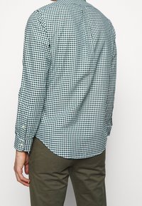 Zelená a bílá kostkovaná košile s dlouhými rukávy a zapínáním na knoflíky na manžetách. Střižená na tělo s mírně zakřiveným spodním lemem a detailem zadního dílu. Nošena s khaki kalhotami.