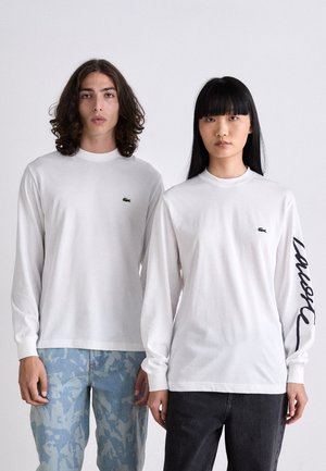Des t-shirts blancs à manches longues présentent un petit logo de crocodile vert sur la poitrine et un texte noir le long de la manche gauche. Coupe décontractée.