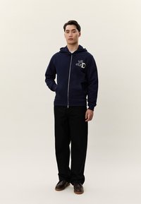 Homem em pé vestindo um capuz azul-marinho com fecho e logótipo "LD", calças pretas e sapatos castanhos contra um fundo bege liso.