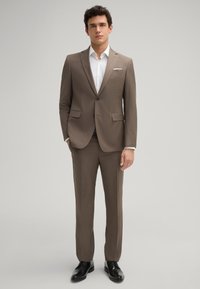 Brauner Maßanzug mit einem Zwei-Knopf-Jacket, Reverskragen und seitlichen Taschen; kombiniert mit einem weißen Hemd und schwarzen Lederschuhen.