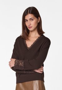 Brauner Pullover mit V-Ausschnitt und Spitzenbesatz am Halsauschnitt und an den Ärmeln. Weiches Material, lässige Passform und filigrane Spitzenverzierungen verleihen Textur.