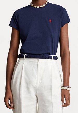 Granatowy bawełniany T-shirt z krótkimi rękawami i okrągłym dekoltem, z małym czerwonym logo. W połączeniu z białymi, wysokim stanem lnianymi spodniami.