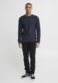 Pulover texturat de nuanță bleumarin cu guler rotund, asociat cu pantaloni negri ajustați și adidași negri cu accente albe.