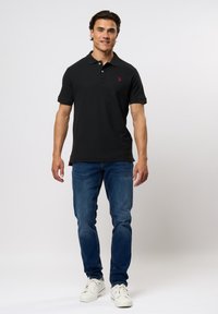 Zwarte polo shirt met korte mouwen, kraag en rood logo. Gecombineerd met blauwe jeans en witte sneakers staat het model tegen een effen achtergrond.