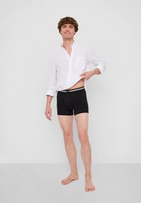 Svarta boxershorts i mjukt tyg, med ett brett midjeband med en kontrasterande rand. Bärs under en vit, oknäppt skjorta.