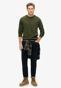 Zielona koszula z długim rękawem, ciemne dżinsy, beżowe buty. Koszula w kratę przewiązana w talii. Prosty design, luźny krój, z minimalnym logo.