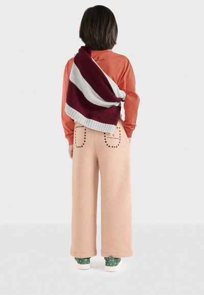 Camicione arancione a maniche lunghe, pantaloni rilassati rosa in pile con tasche a pois e un maglione lavorato a maglia grigio e rosso a righe appoggiato sulla spalla.