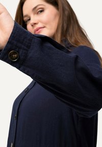 Marineblauw overhemdd met lange mouwen, met een zichtbare bruine knoopdetail en een zachte textuur. De stof heeft een lichte glans.