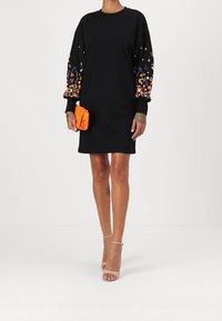 Svart sweatshirtklänning med blommig broderi på ärmarna, med en lös passform. Tillsammans med en texturerad orange kuvertväska och beigefärgade klackar.