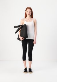 Tilpasset hvit tanktop, svarte korte leggings og svarte sko med gummisåler. Modellen holder en svart veske med store håndtak. Enfarget bakgrunn.