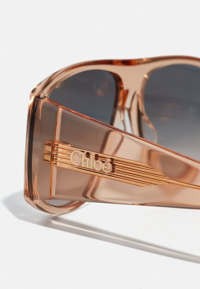 Ochelari de soare supradimensionați cu un gradient roz, cadru transparent și lentile închise la culoare. Prezintă un logo „Chloé” auriu pe un braț texturat.