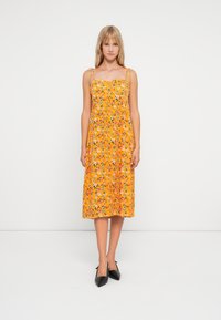 Vestido amarelo florido com alças finas, apresentando uma cintura franzida e saia de comprimento médio. Sapatos de salto alto pretos completam o conjunto.