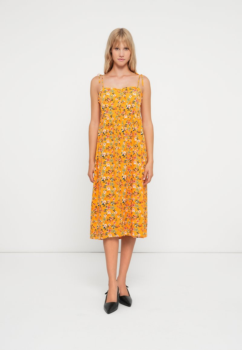 Vestido amarelo florido com alças finas, apresentando uma cintura franzida e saia de comprimento médio. Sapatos de salto alto pretos completam o conjunto.