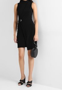 Černé šaty bez rukávů s detailem postranního uzlu a kruhovým kovovým akcentem. Kombinovány s černými mokasínami na podpatku a kulatou kabelkou.