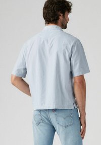 Camicia a maniche corte con colletto, di colore azzurro chiaro e righe verticali. Realizzata in tessuto morbido; presenta un taglio dritto e una spalla sul retro.